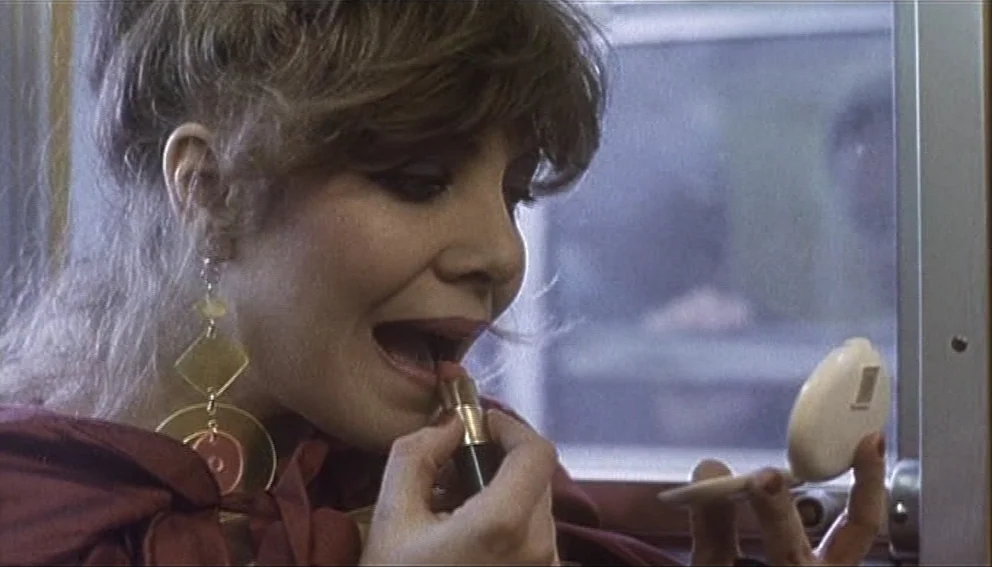 Labyrinth of Passion (1982) movie still: closeup — A woman sits inside a subway car with windows visible in the background. She is applying lipstick…