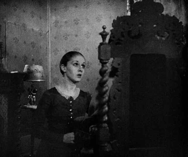 Vampyr (1932) movie still: medium — A dimly lit room features patterned wallpaper and an ornate carved wooden chair back. A woman stands in the…