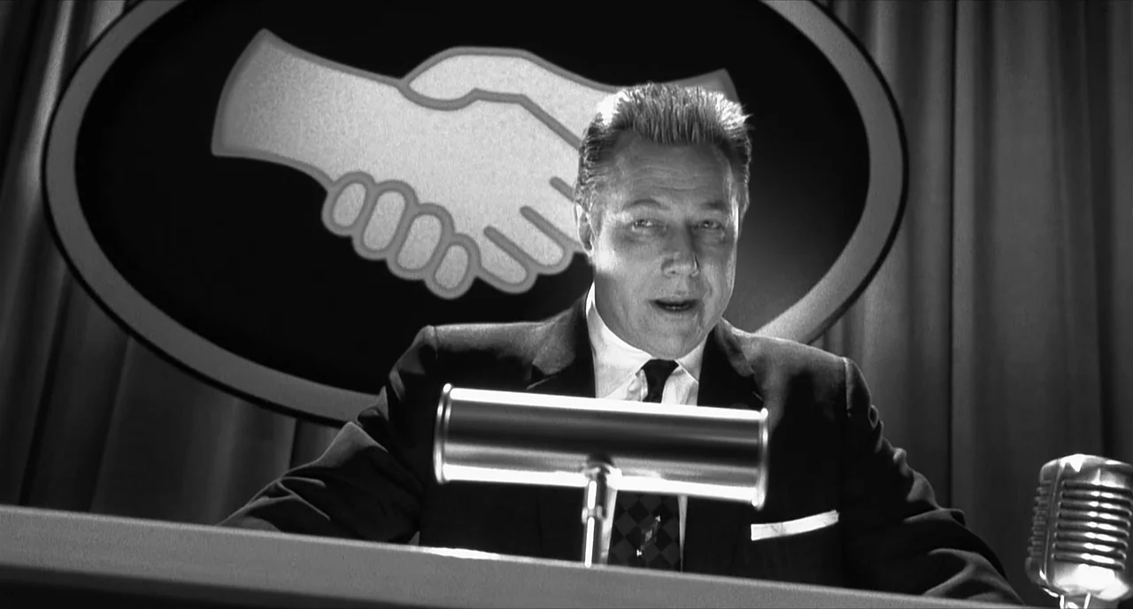 Pleasantville (1998) movie still: medium — A television desk set features a large oval handshake logo on the back wall. A man in a suit sits at the…
