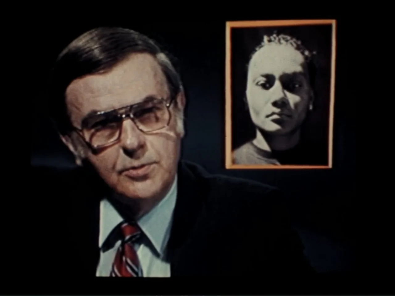 Born in Flames (1983) movie still: medium — A television studio environment features a dark, unlit background behind a single male subject. The man…