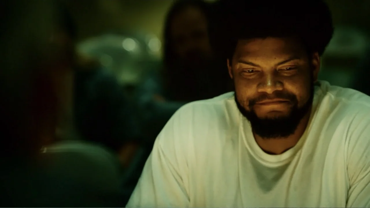 Hancock (2008) movie still: medium — A dimly lit indoor space with blurred figures in the background. A man sits facing forward in a white shirt…