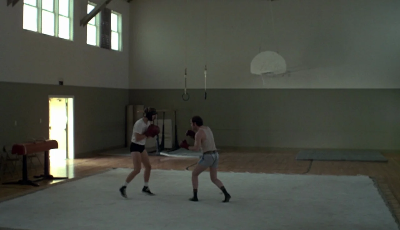 Fat City (1972) movie still: wide — A large, sparse gymnasium with wooden floors and tall windows lining the upper wall. Two men in boxing gear face…
