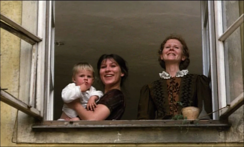 Woyzeck (1979) movie still: medium — A group of three people look out from a square window set into a wall. The two women and a small child smile…