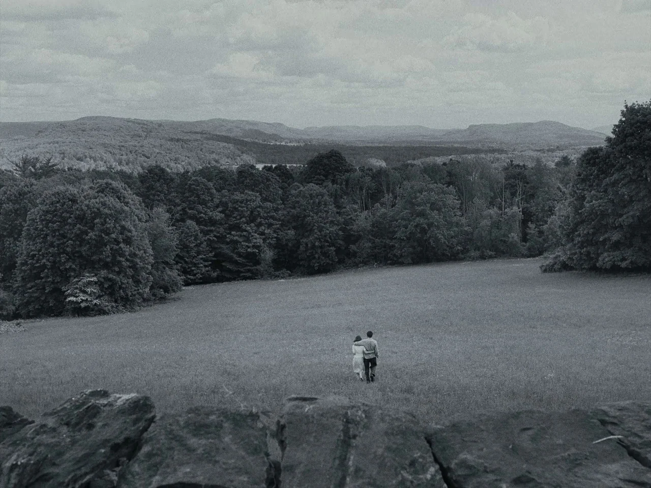 Maestro (2023) movie still: wide — A vast grassy meadow sits in front of a dense forest and rolling mountains. A young couple walks across the…