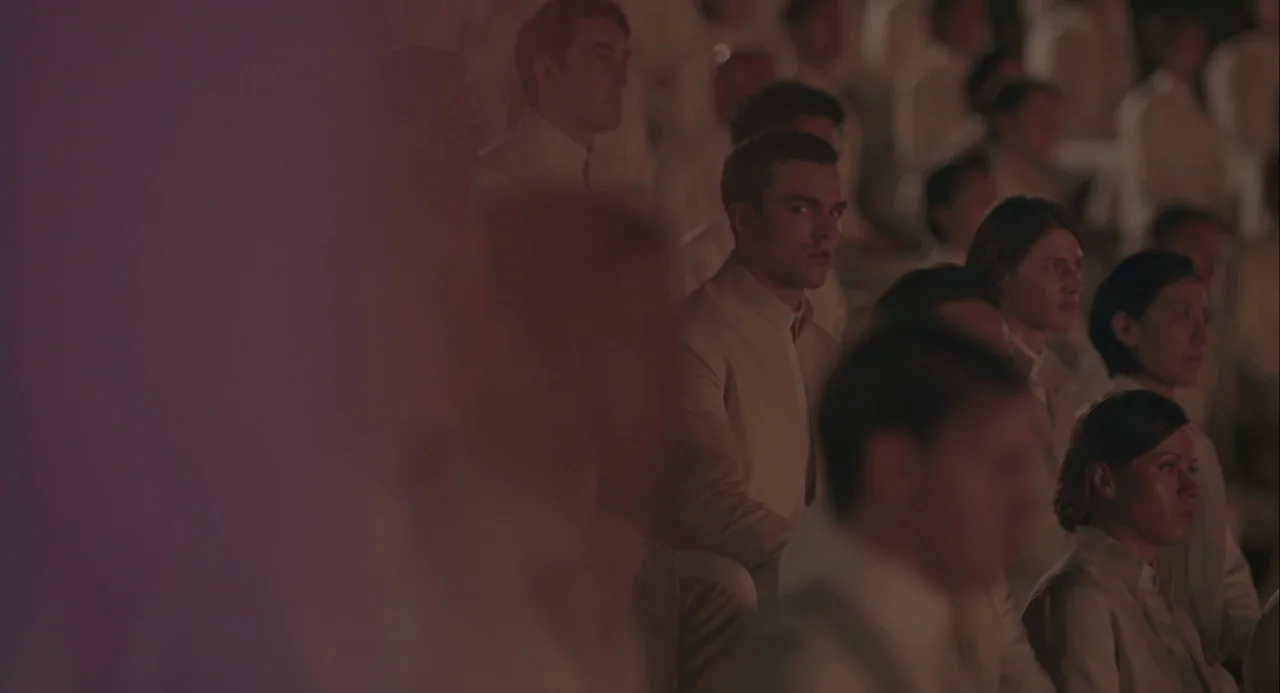 Equals (2015) movie still: medium — A dimly lit auditorium filled with rows of people wearing uniform pale beige clothing. A young man sits among…
