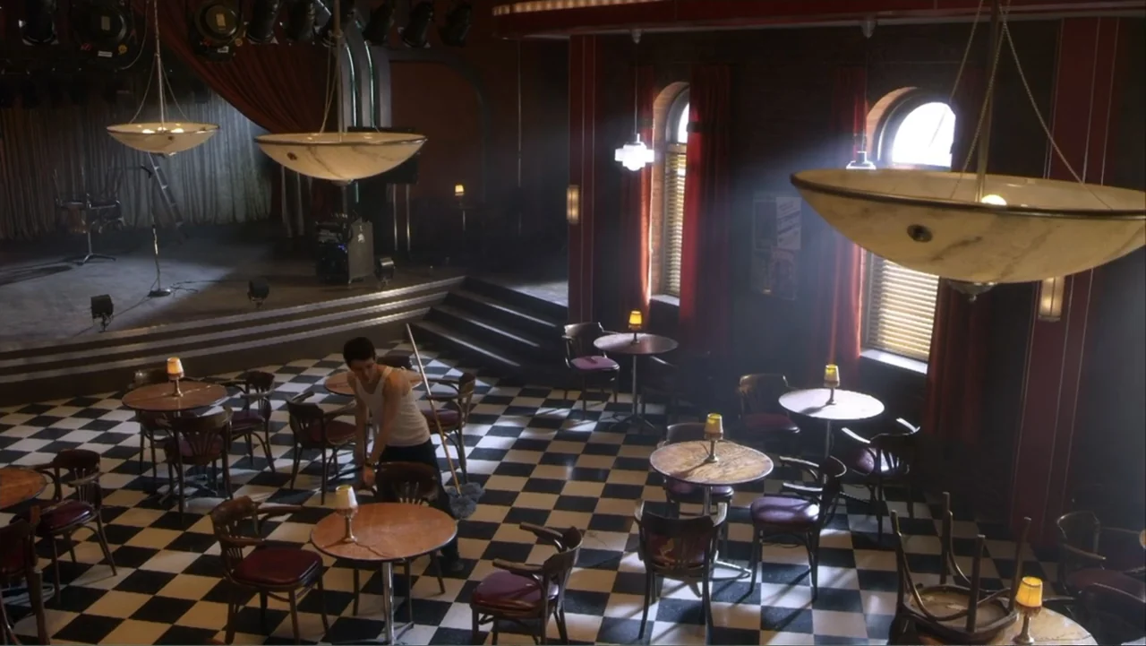 Rags (2012) movie still: wide — A spacious restaurant interior features a black and white checkered floor and round tables. A man wearing a white…