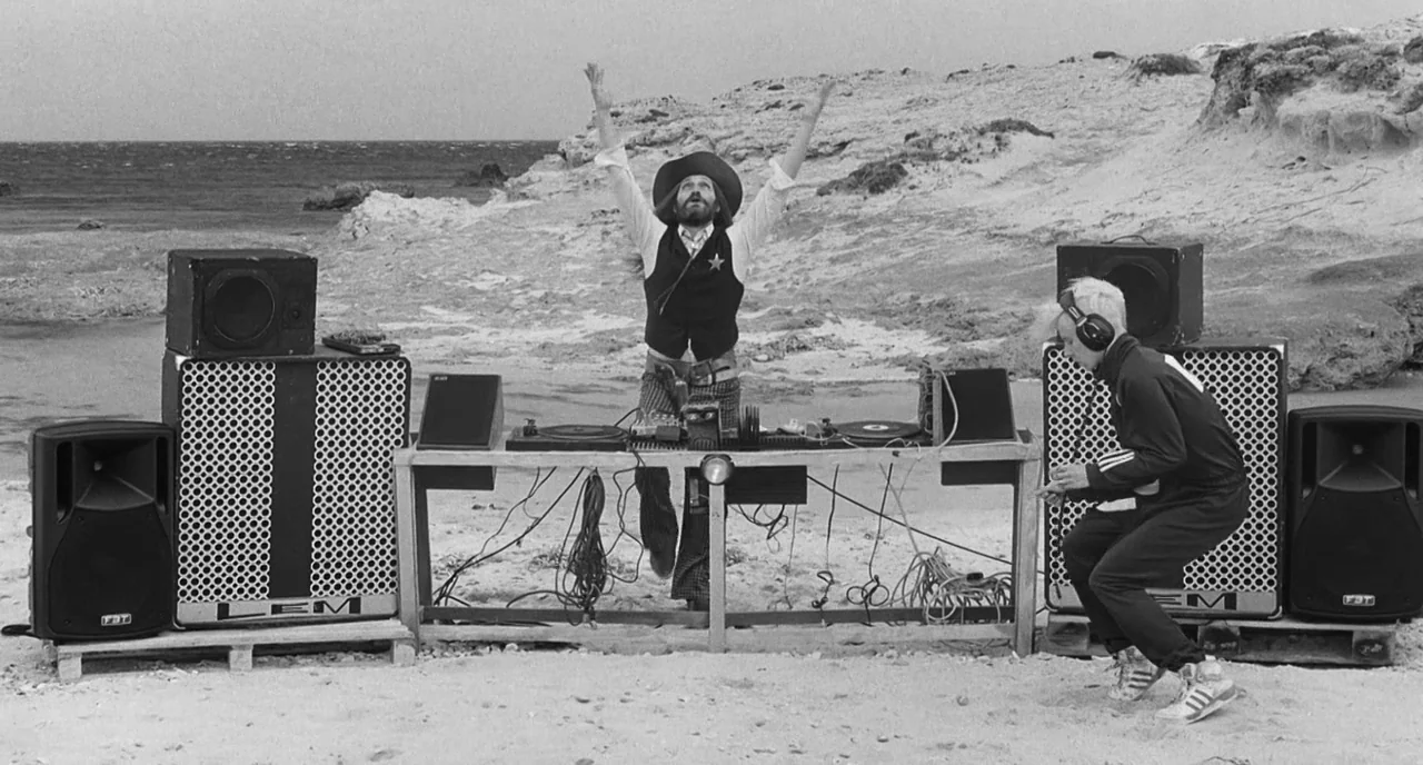 The Legend Of Kaspar Hauser (2012) movie still: wide — A DJ setup on a sandy beach featuring large speakers and electronic audio equipment. One man…
