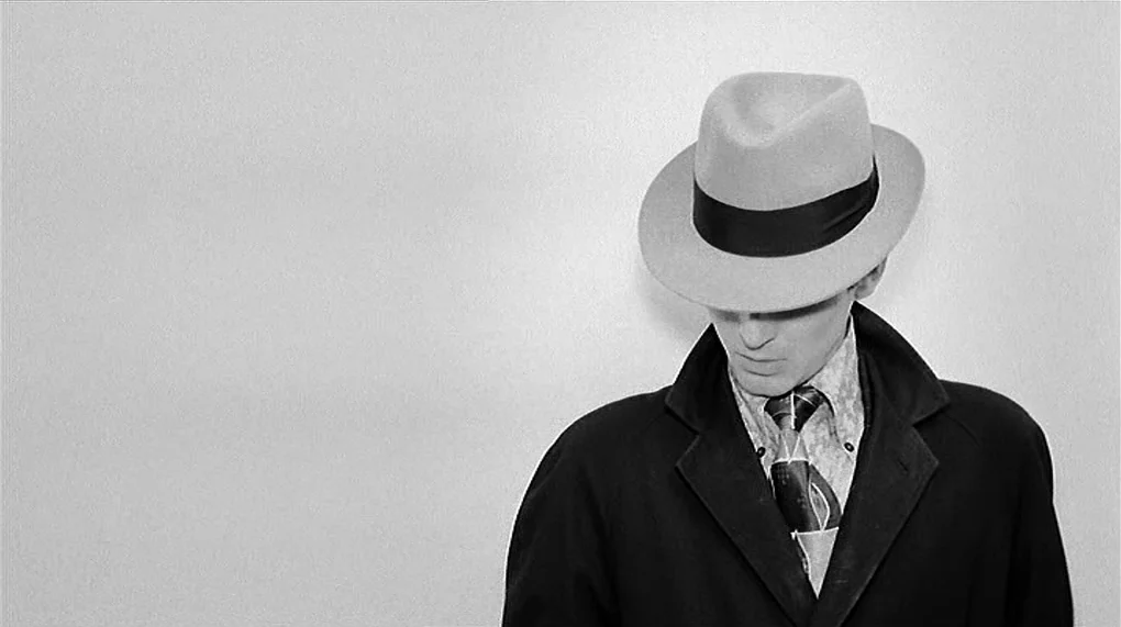 Love Is Colder Than Death (1969) movie still: medium — A man stands against a plain white wall in a sterile indoor setting. He wears a fedora hat…