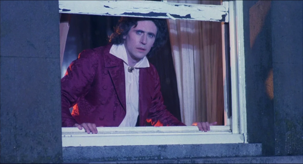 Gothic (1986) movie still: A striking medium-closeup shot frames a young man in an ornate maroon jacket as he leans out of a dilapidated window,…