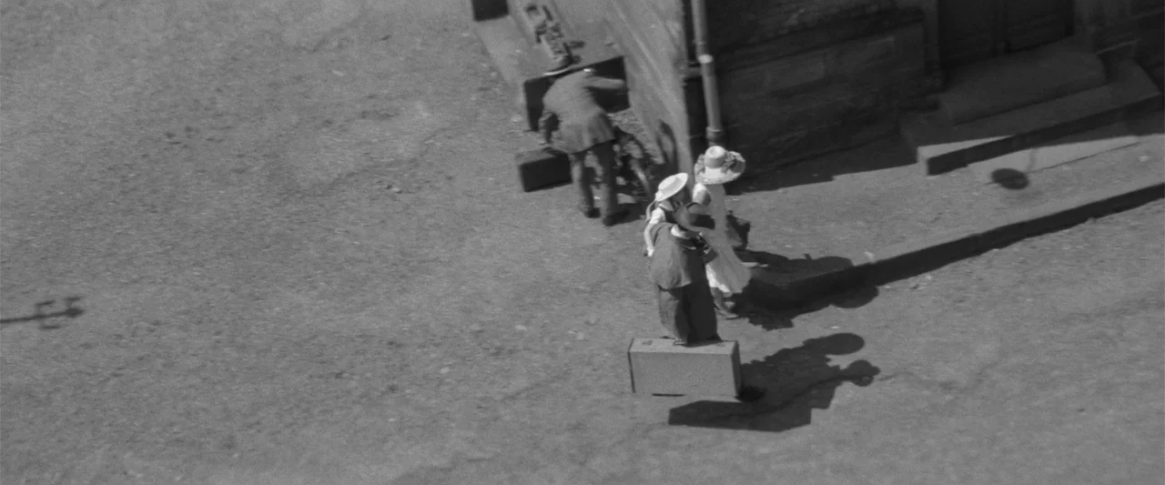 Jules and Jim (1962) movie still: wide — A group of people dressed in 1920s attire walk on a paved outdoor area. They carry suitcases while moving…
