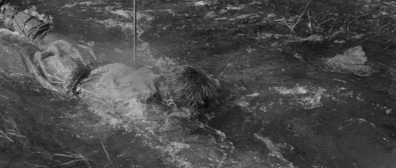 Andrei Rublev (1966) movie still: medium — A person lies submerged in a shallow, fast-moving stream with turbulent water. The subject is oriented…