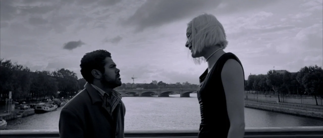 Angel-A (2005) movie still: medium — A stone bridge overlooking a river under a cloudy sky in a city. A man and a blonde woman stand facing each…