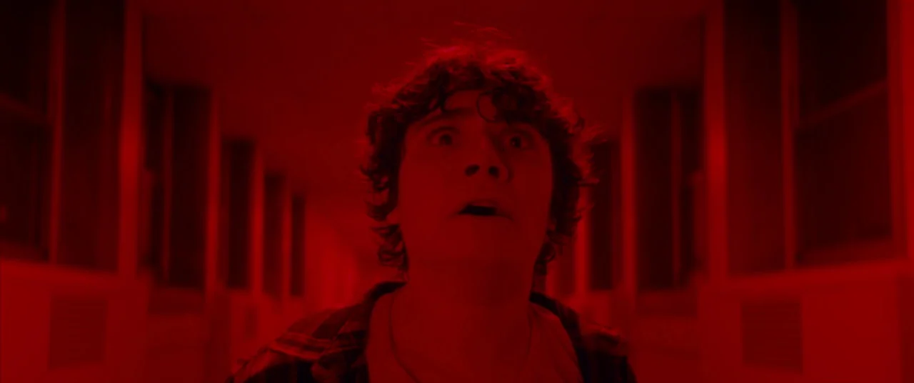 Scary Stories to Tell in the Dark (2019) movie still: closeup — A long, dimly lit indoor corridor bathed in intense, saturated red light. A young…
