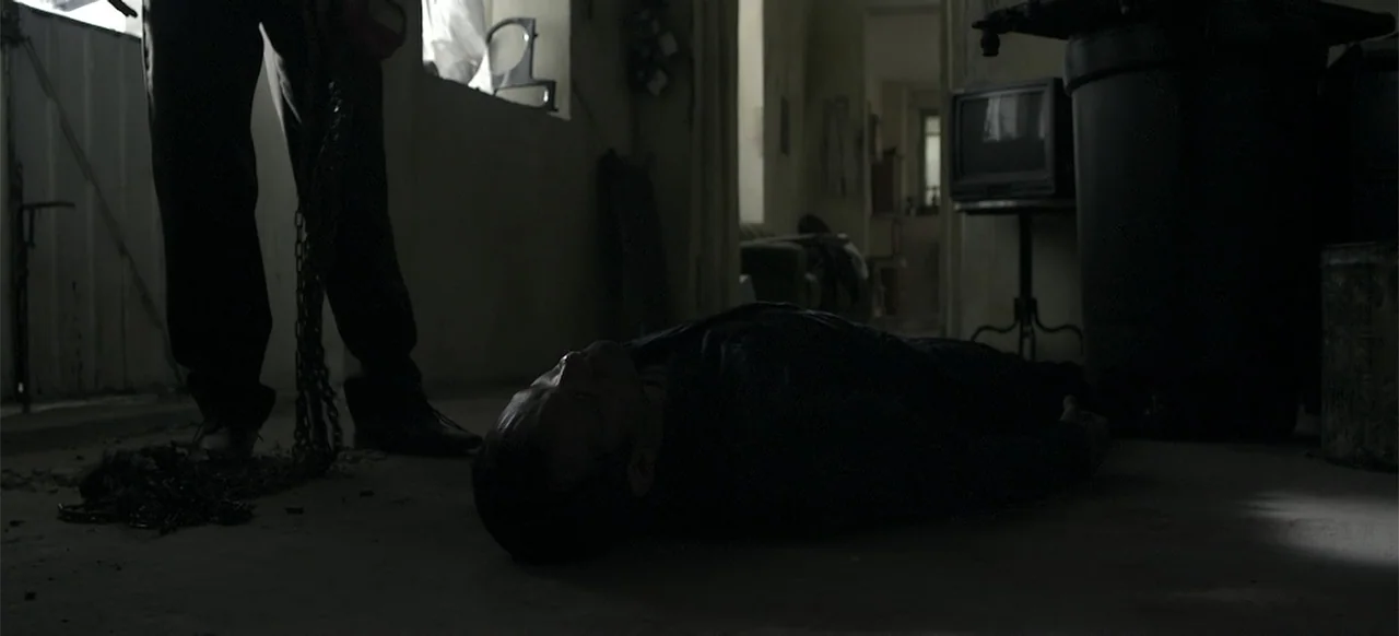About the Deaths (2016) movie still: medium — A dimly lit room with peeling paint on the walls and a television nearby. A man lies prone on the…