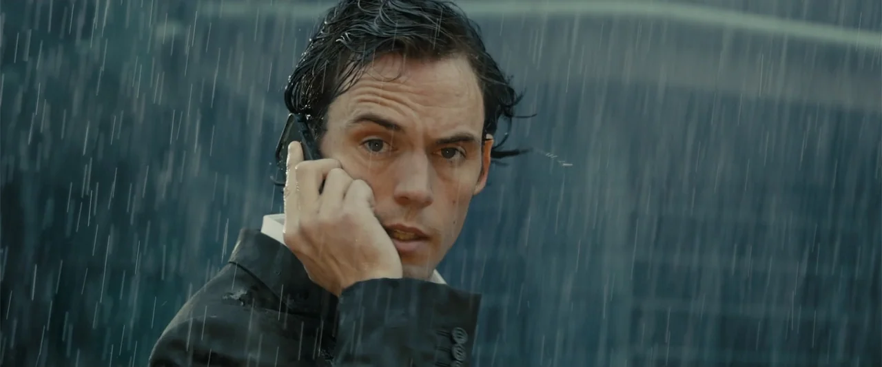 Me Before You (2016) movie still: closeup — A man stands outdoors in the rain against a blurred building background. He is holding a mobile phone to…
