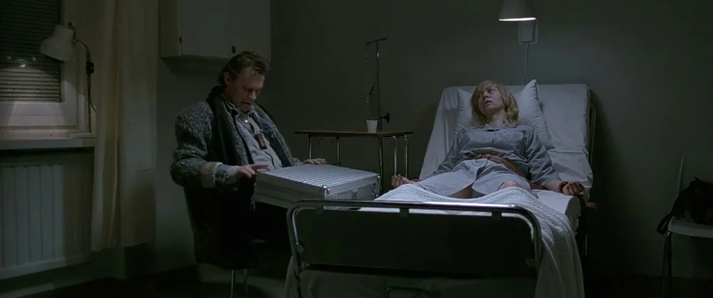 Let The Right One In (2008) movie still: wide — A dim, stark hospital room features a metal bed occupied by a resting patient. A man sitting in a…