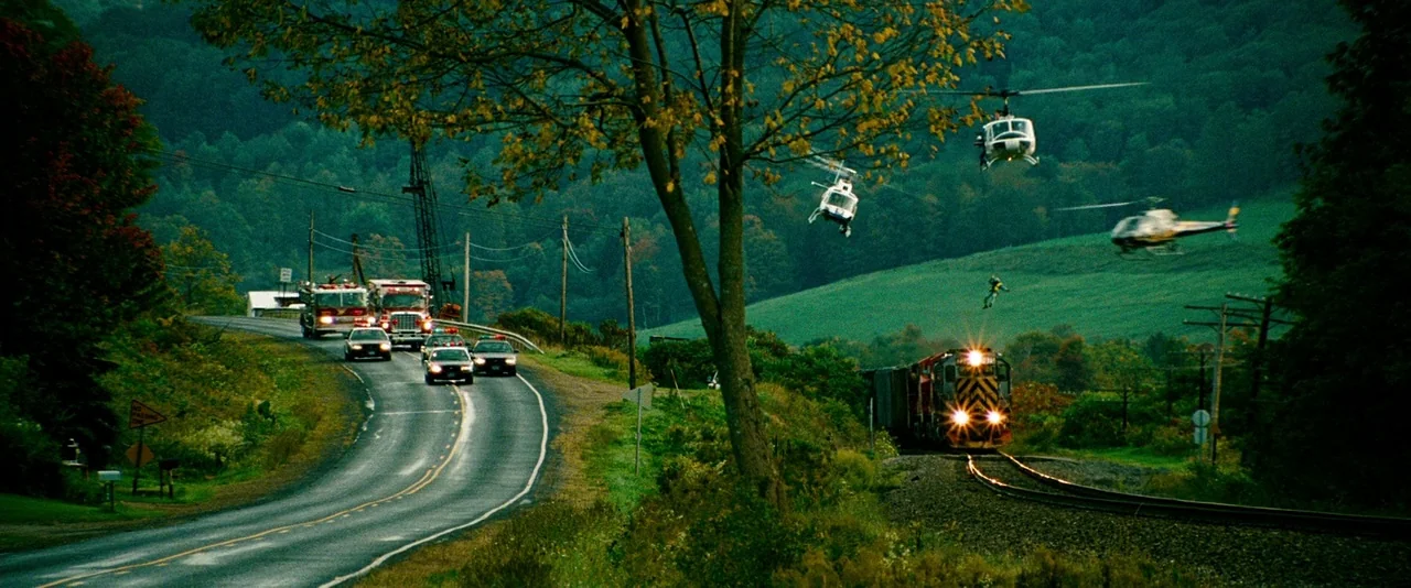 Unstoppable (2010) movie still: wide — A rural landscape features a winding road alongside train tracks and dense trees. A locomotive travels along…