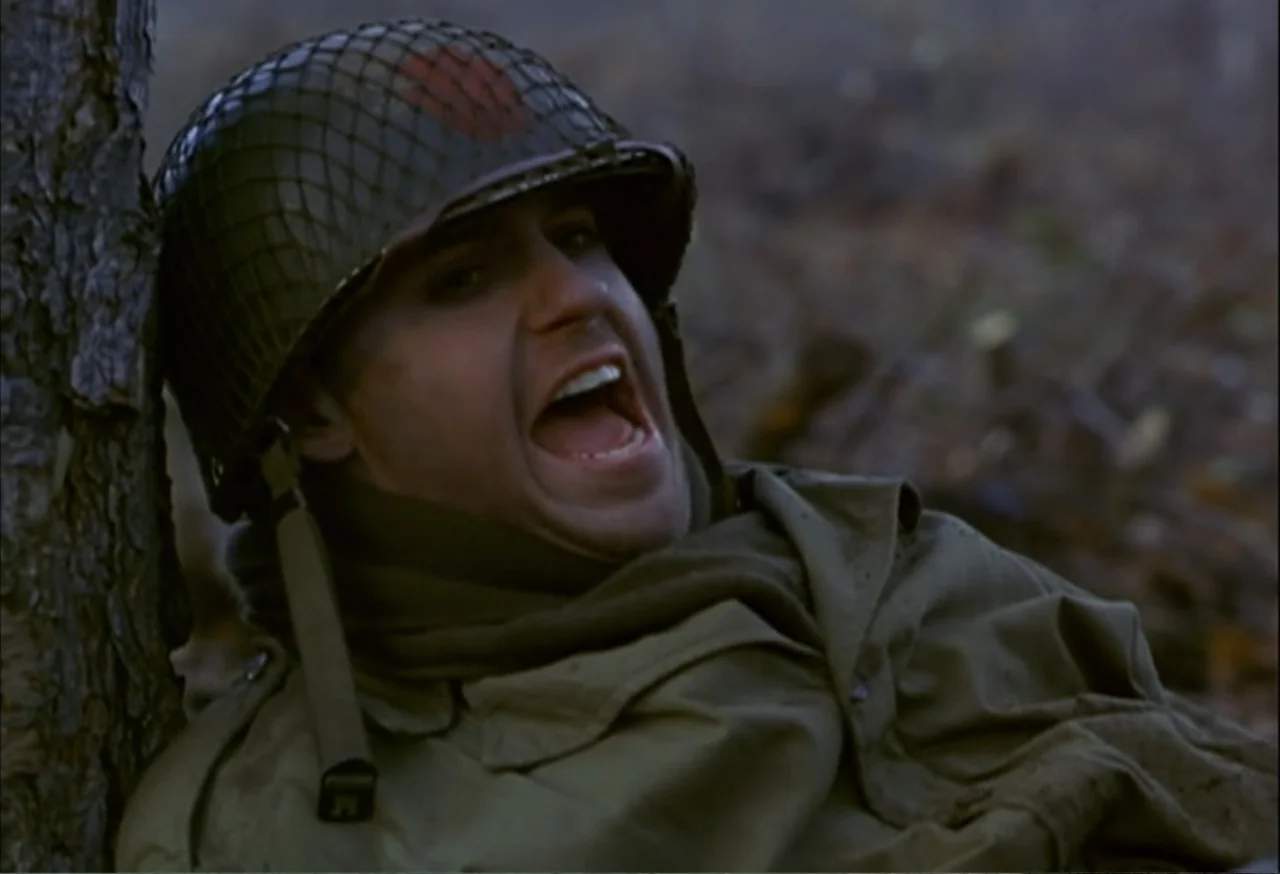 When Trumpets Fade (1998) movie still: closeup — A soldier in uniform rests against a tree trunk in a forested environment. The man is shouting with…