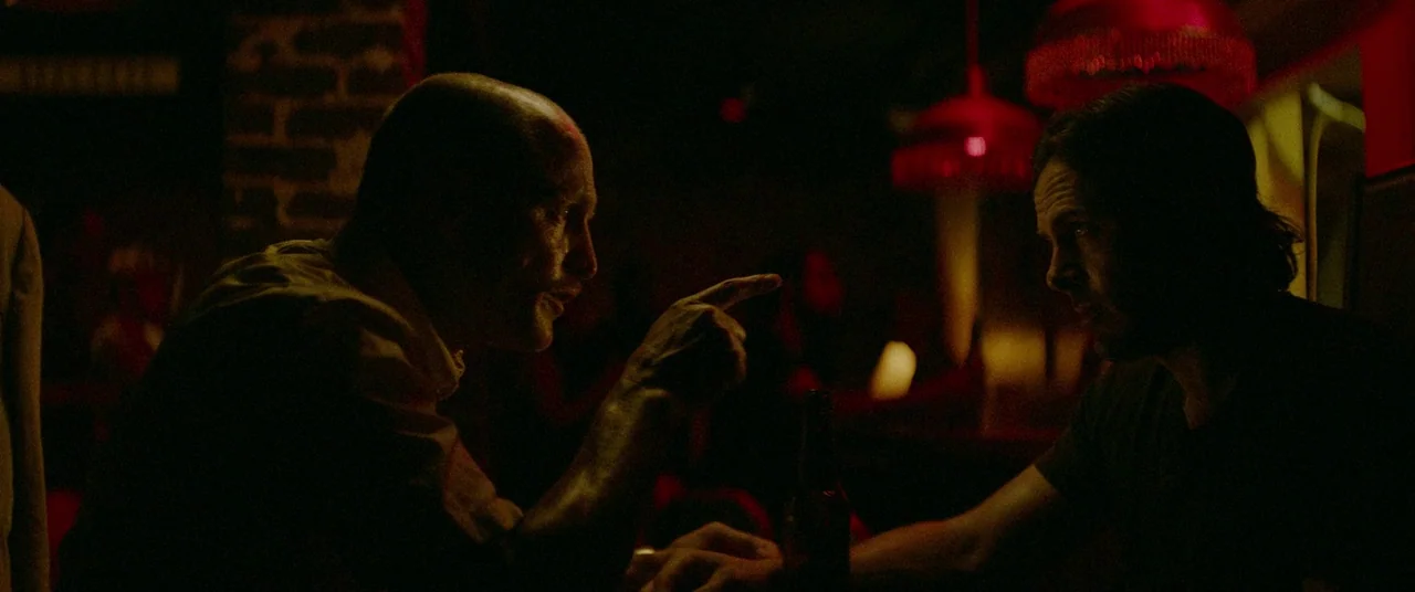 Triple 9 (2016) movie still: medium — A dimly lit bar space with red hanging lights and exposed brick textures. Two men sit at a table facing each…