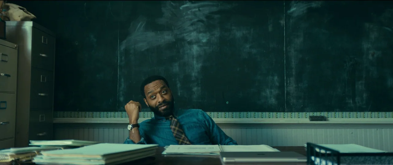 The Life of Chuck (2024) movie still: medium — A dark, chalk-covered blackboard serves as the background in an empty classroom setting. A man in a…