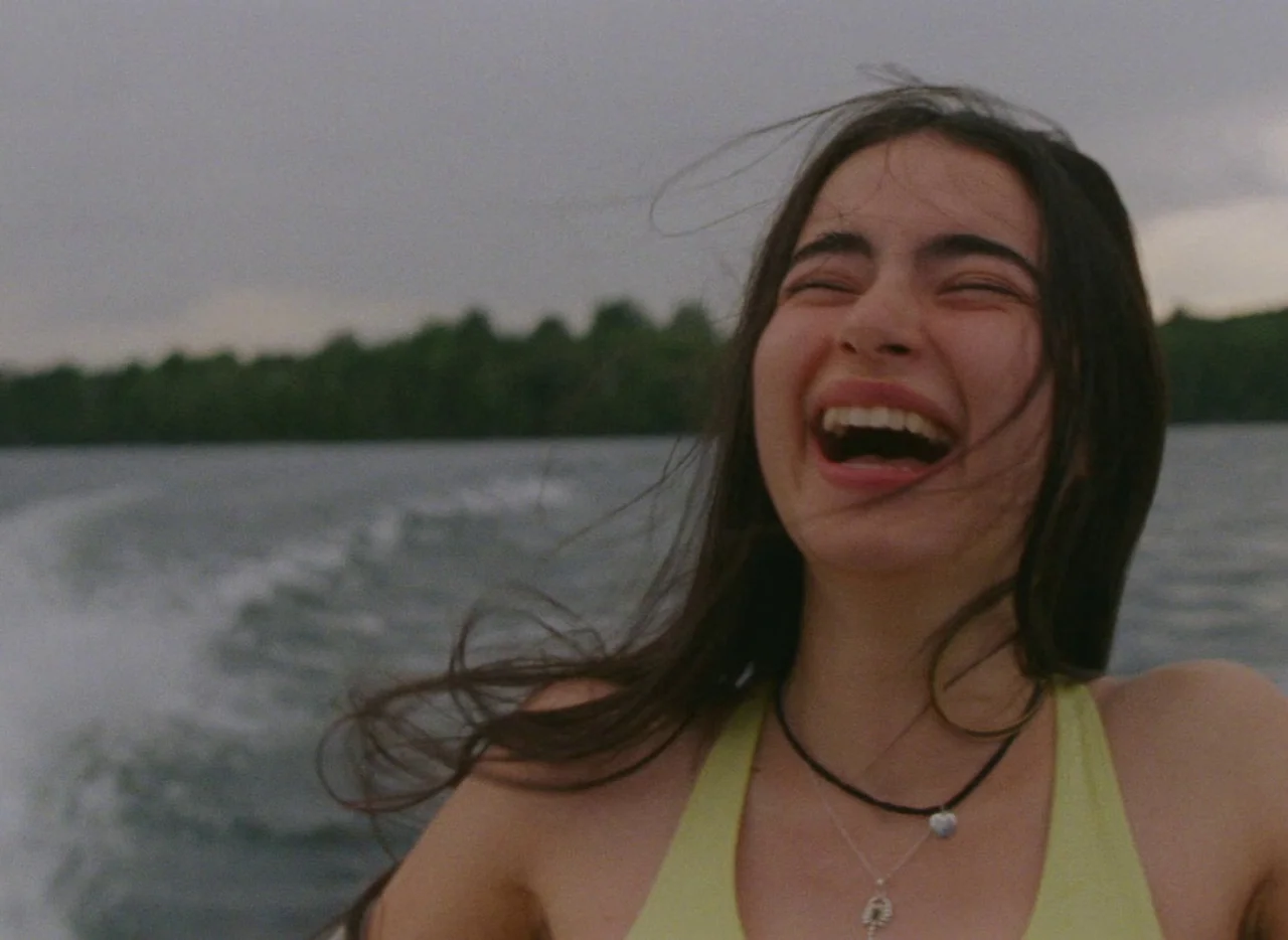 Falcon Lake (2022) movie still: closeup — A woman laughs while sitting on a moving boat on a large body of water. Distant dark green treeline is…