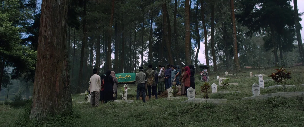 Satan’s Slaves (2017) movie still: wide — A group of mourners stands around a green casket in a sloping hillside cemetery. Tall pine trees surround…