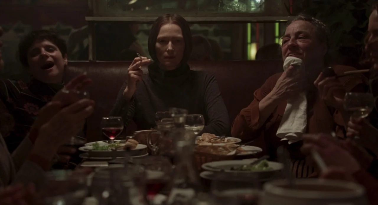 Suspiria (2018) movie still: medium — A group of people sits around a dark table filled with plates and glasses. The woman in the center holds a…