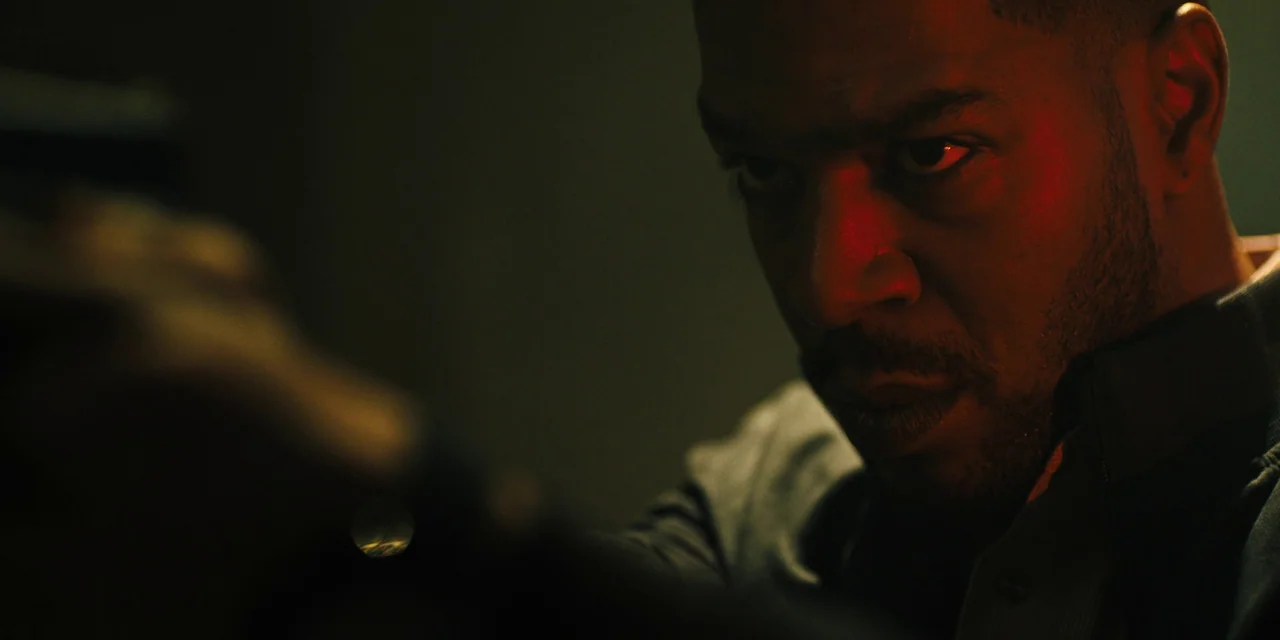Silent Night (2023) movie still: closeup — A man stands in a dark, poorly lit space with limited visibility. He aims a firearm directly toward the…