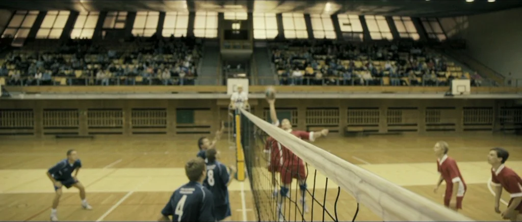 Lasting (Nieulotne) (2013) movie still: wide — An indoor volleyball court features a net dividing two teams of athletes wearing uniforms. A crowd…
