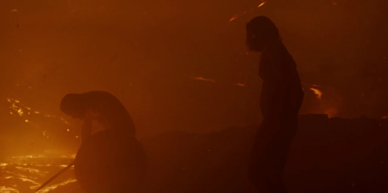 The Northman (2022) movie still: wide — A rocky volcanic surface features flowing molten rock amidst deep orange atmosphere. Two figures appear in…