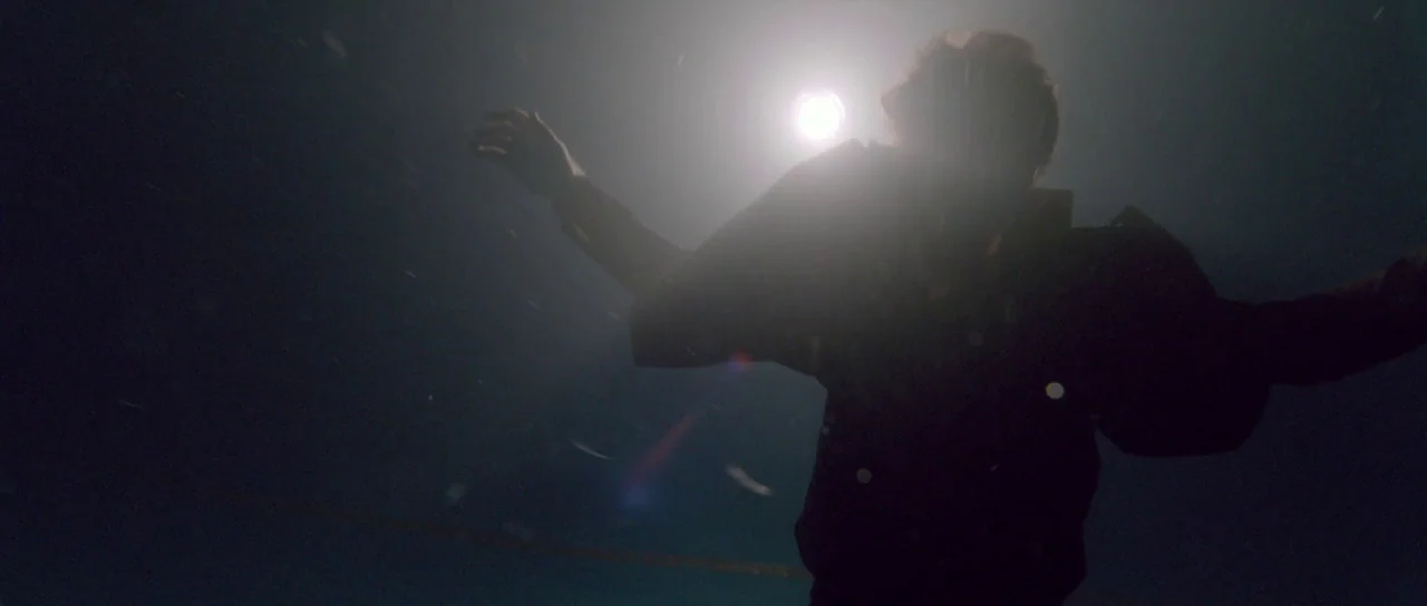Knight of Cups (2015) movie still: medium — A person drifts in dark water beneath a bright light source at night. Floating particles are visible in…
