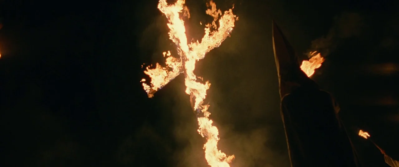 BlacKkKlansman (2018) movie still: wide — A dark, open field at night features a large, upright wooden cross engulfed in flames. A person wearing a…