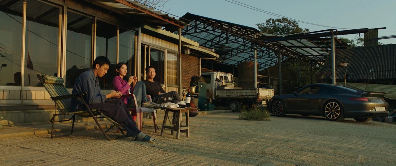 Burning (2018) movie still: wide — A rural yard with a weathered house and an adjacent metal-roofed agricultural structure. Three individuals sit…