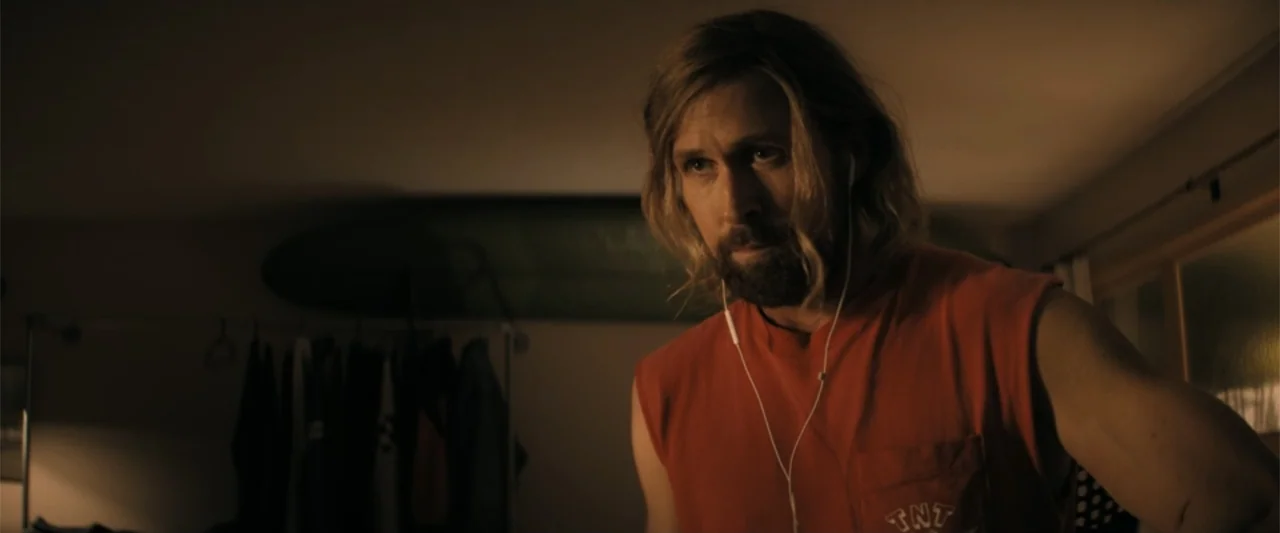 The Fall Guy (2024) movie still: medium — A dimly lit room features a surfboard mounted on a wall behind a rack. A bearded man in a red tank top…