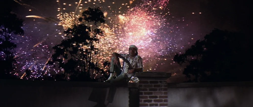 Cape Fear (1991) movie still: fullbody — A man sits alone on a low brick wall at night under a dark sky. A massive fireworks display erupts in…