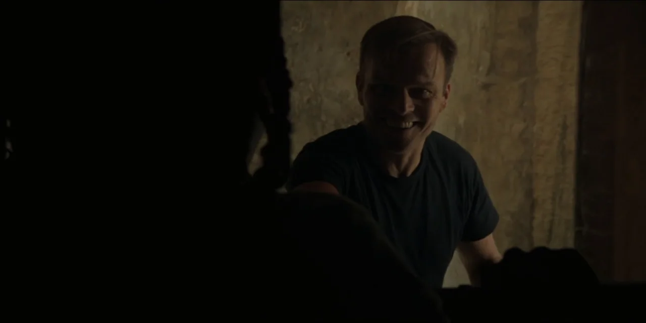 The Hunted (2024) movie still: medium — A dimly lit indoor space features textured walls and a narrow passageway. A man in a dark shirt smiles at an…