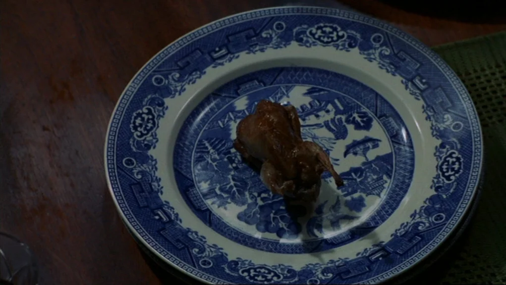 Frenzy (1972) movie still: closeup — A small roasted quail sits on a blue and white patterned willow ware plate. The plate is positioned on a dark…