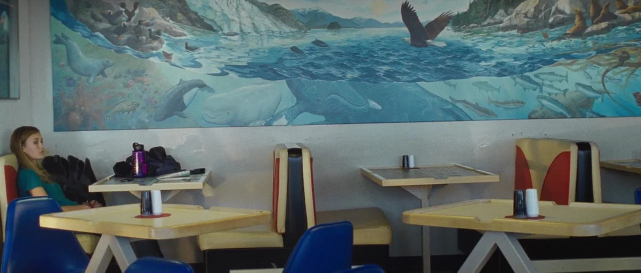 Wildlike (2014) movie still: wide — A diner booth area features a large panoramic mural depicting coastal wildlife on the wall. A woman sits alone…