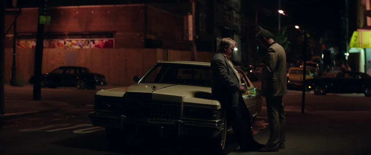 God’s Pocket (2014) movie still: wide — A night scene on a city street with dark buildings and a white car. Two men stand beside the vehicle engaged…