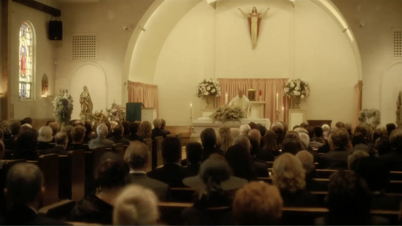 Behind the Candelabra (2013) movie still: wide — A crowded funeral chapel features rows of wooden pews facing a central coffin. An officiant stands…