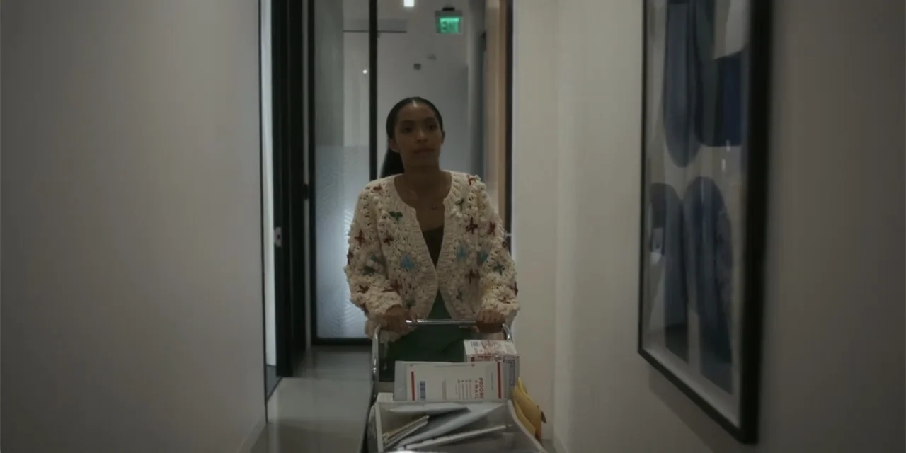Sitting in Bars with Cake (2023) movie still: medium — A modern office hallway with white walls and a framed abstract blue print hanging. A young…