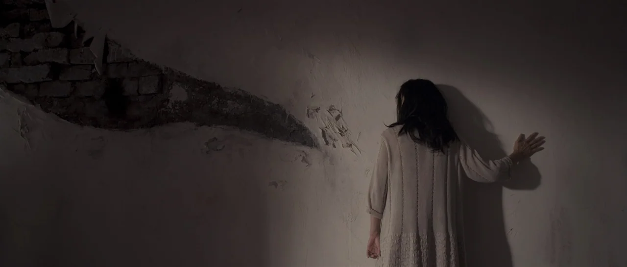 Rigor Mortis (2013) movie still: medium — A dimly lit room features a white wall with a large, jagged patch of exposed brick. A woman stands with…