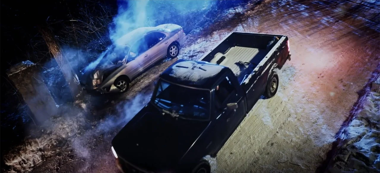 The Russian Bride (2019) movie still: wide — A snow-covered dirt road at night shows a wrecked sedan emitting plumes of smoke. A dark pickup truck…