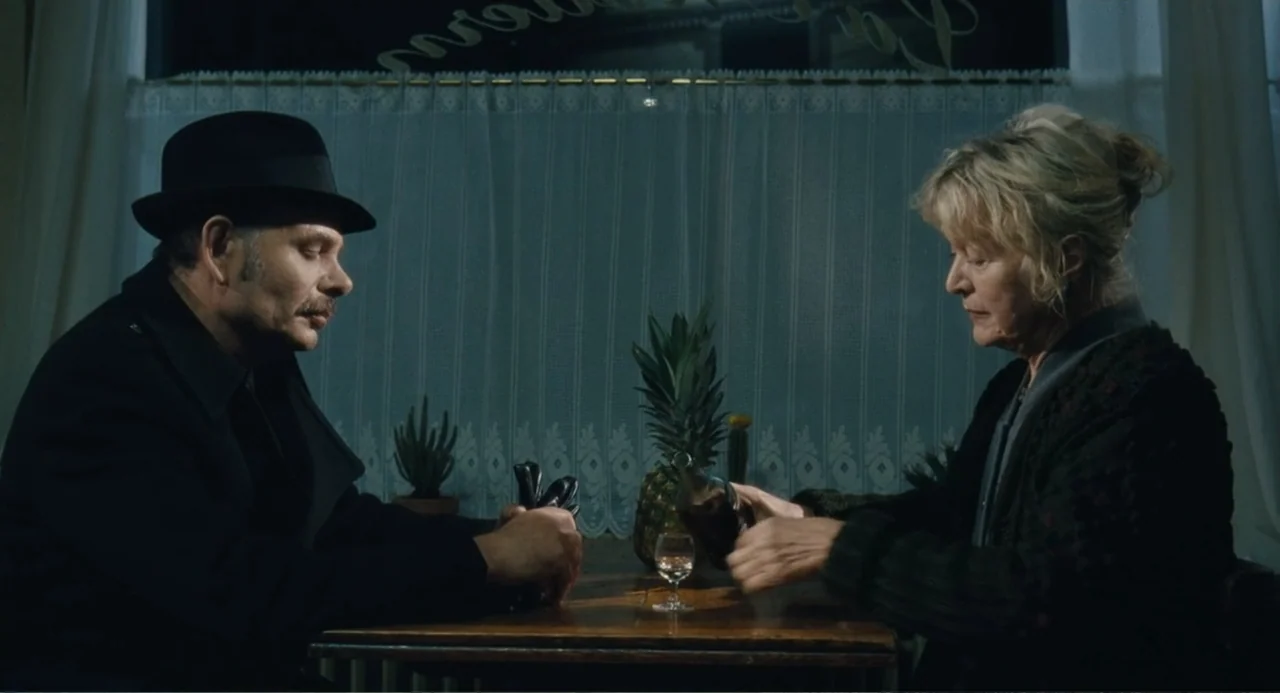 Le Havre (2011) movie still: medium — A dimly lit cafe interior with a lace curtain covering the window at night. A man in a hat and an older woman…