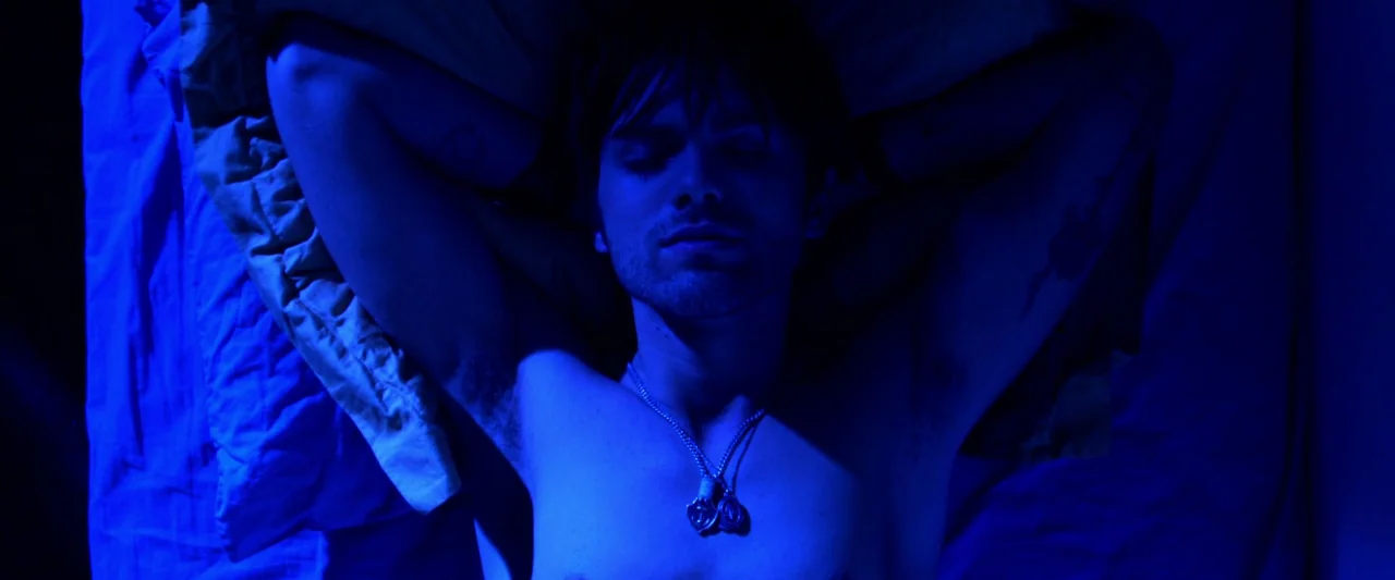 Kaboom (2010) movie still: closeup — A person lies on a mattress covered in white sheets. The subject rests with arms folded behind their head.