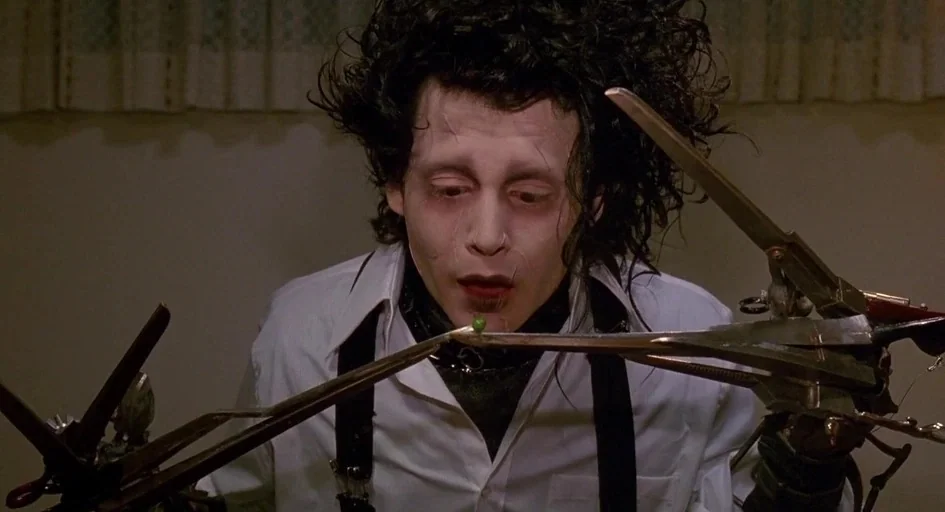 Edward Scissorhands (1990) movie still: closeup — A man with scissor-hands sits in a domestic dining room with cream-colored walls. He carefully…