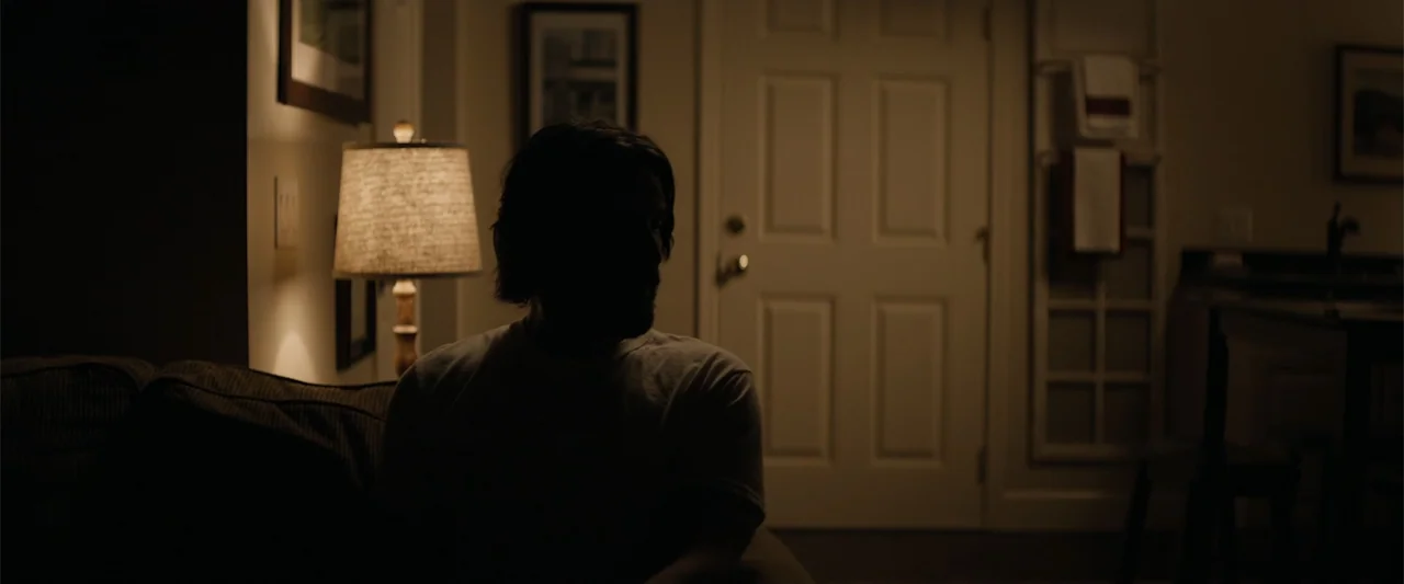 Destination Wedding (2018) movie still: medium — A dimly lit room features a sofa, a table lamp, and a closed door. A man sits on the sofa with his…