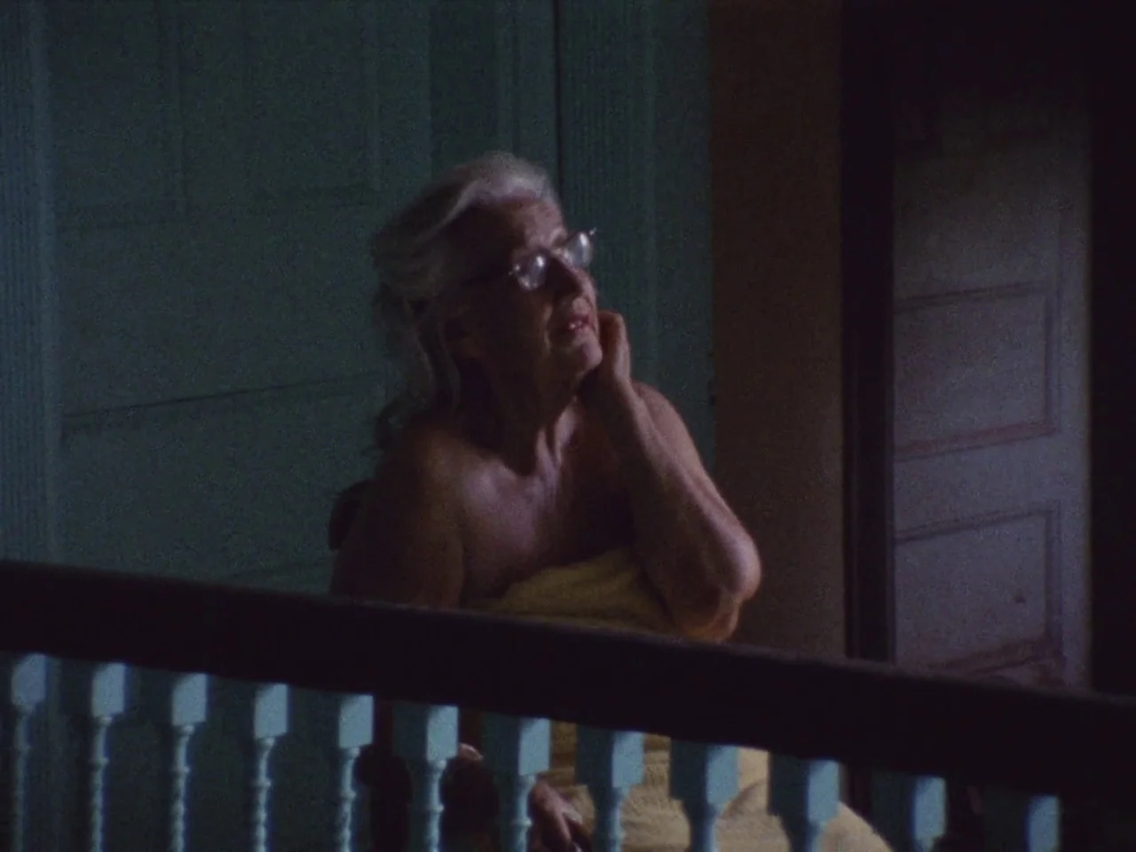 Grey Gardens (1975) movie still: medium — A dimly lit staircase landing features a wooden balustrade and shadowed walls. An elderly woman in a towel…