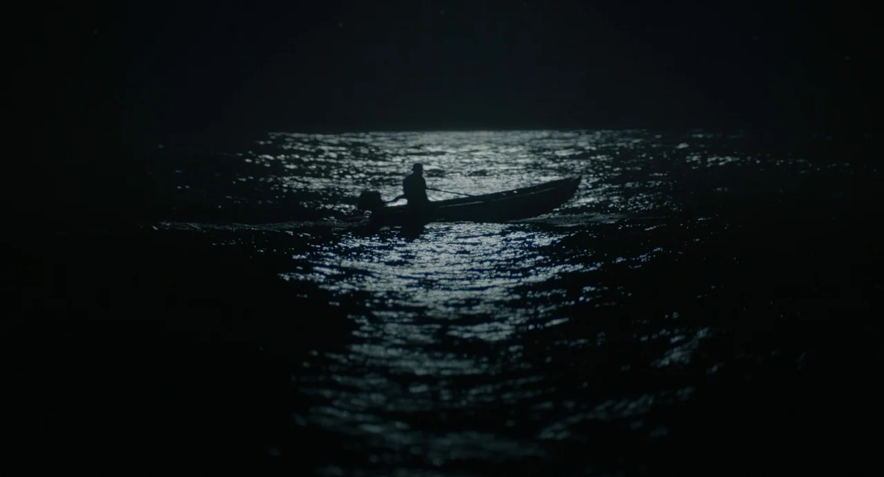 Beau Is Afraid (2023) movie still: wide — A lone person rows a small boat across a dark, rippling body of water. Moonlight highlights the surface of…