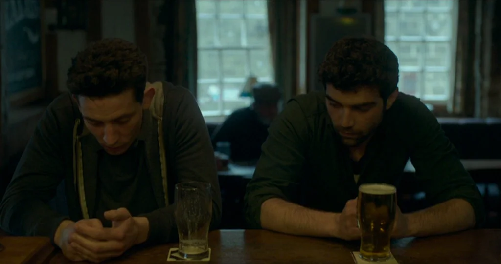 God’s Own Country (2017) movie still: medium — A rustic interior of a dimly lit local pub with paneled walls and windows. Two men sit at the wooden…